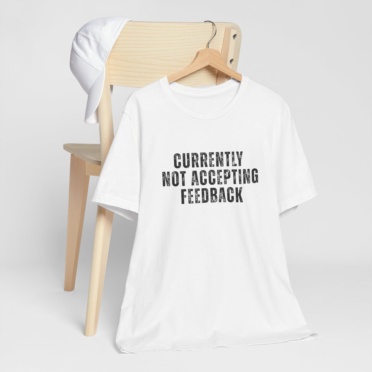 A white short-sleeve t-shirt with the text 'CURRENTLY NOT ACCEPTING FEEDBACK' printed in black distressed capital letters, hanging on a wooden hanger in front of a light wood chair with a white cap resting on it, against a light gray background.